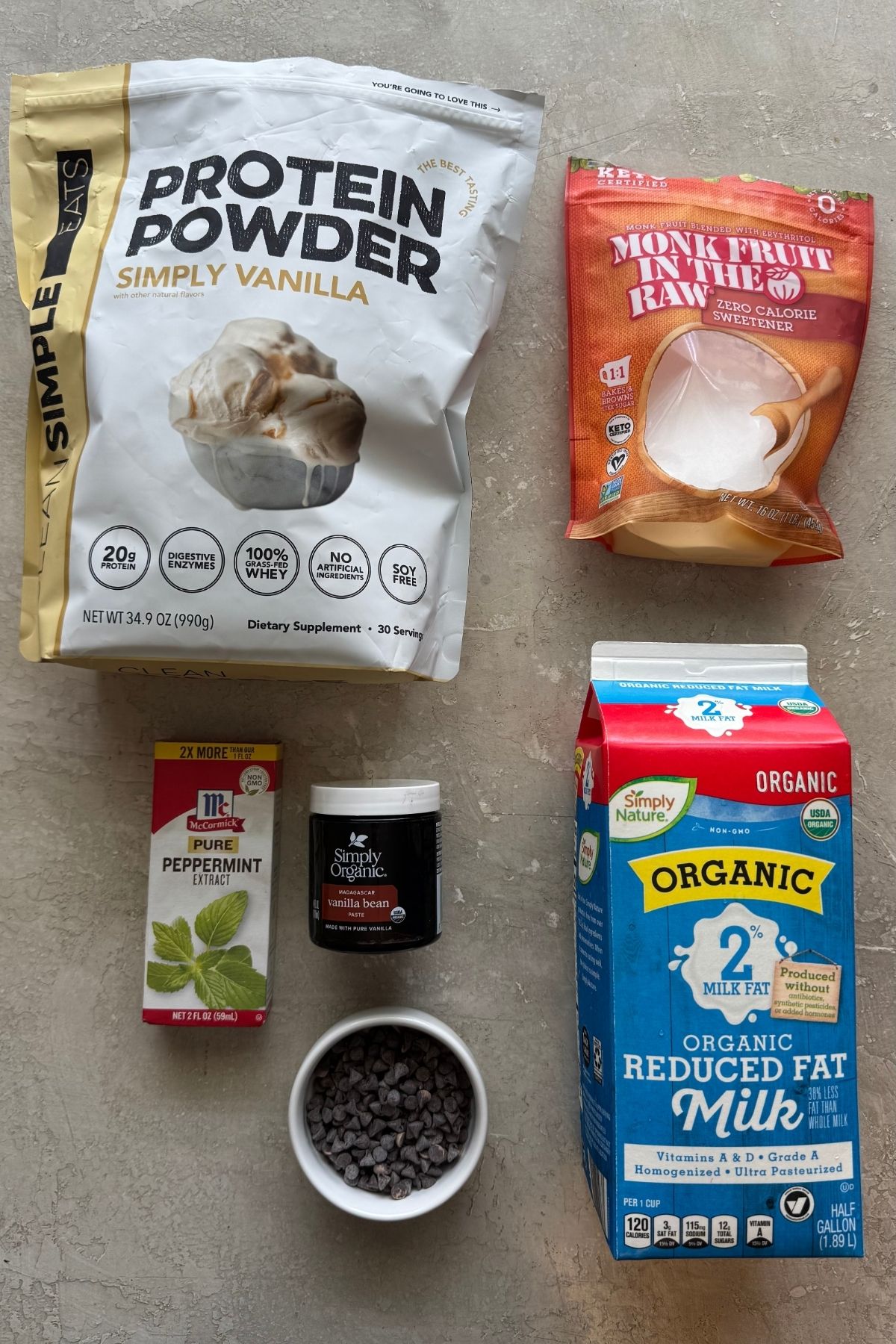 Flat lay of ingredients for Mint Chocolate Protein Ice Cream: Simply Vanilla protein powder, Monk Fruit sweetener, McCormick peppermint extract, vanilla bean paste, chocolate chips, and organic milk on a grey surface.