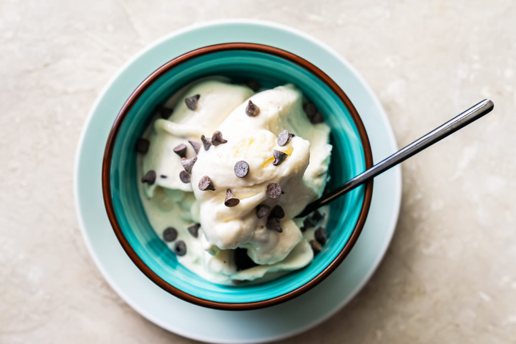 Overhead view of a teal bowl filled with Mint Chocolate Protein Ice Cream and chocolate chips, resting on a matching light blue saucer. A silver spoon is inserted into the side of the creamy white dessert.