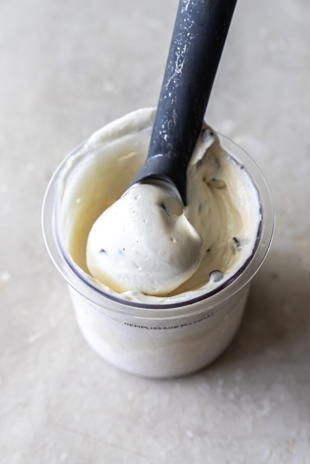 Close-up of a black ice cream scoop lifting a ball of white Mint Chocolate Protein Ice Cream from a clear Ninja Creami pint. Small chocolate chips are visible within the creamy base against a light background.
