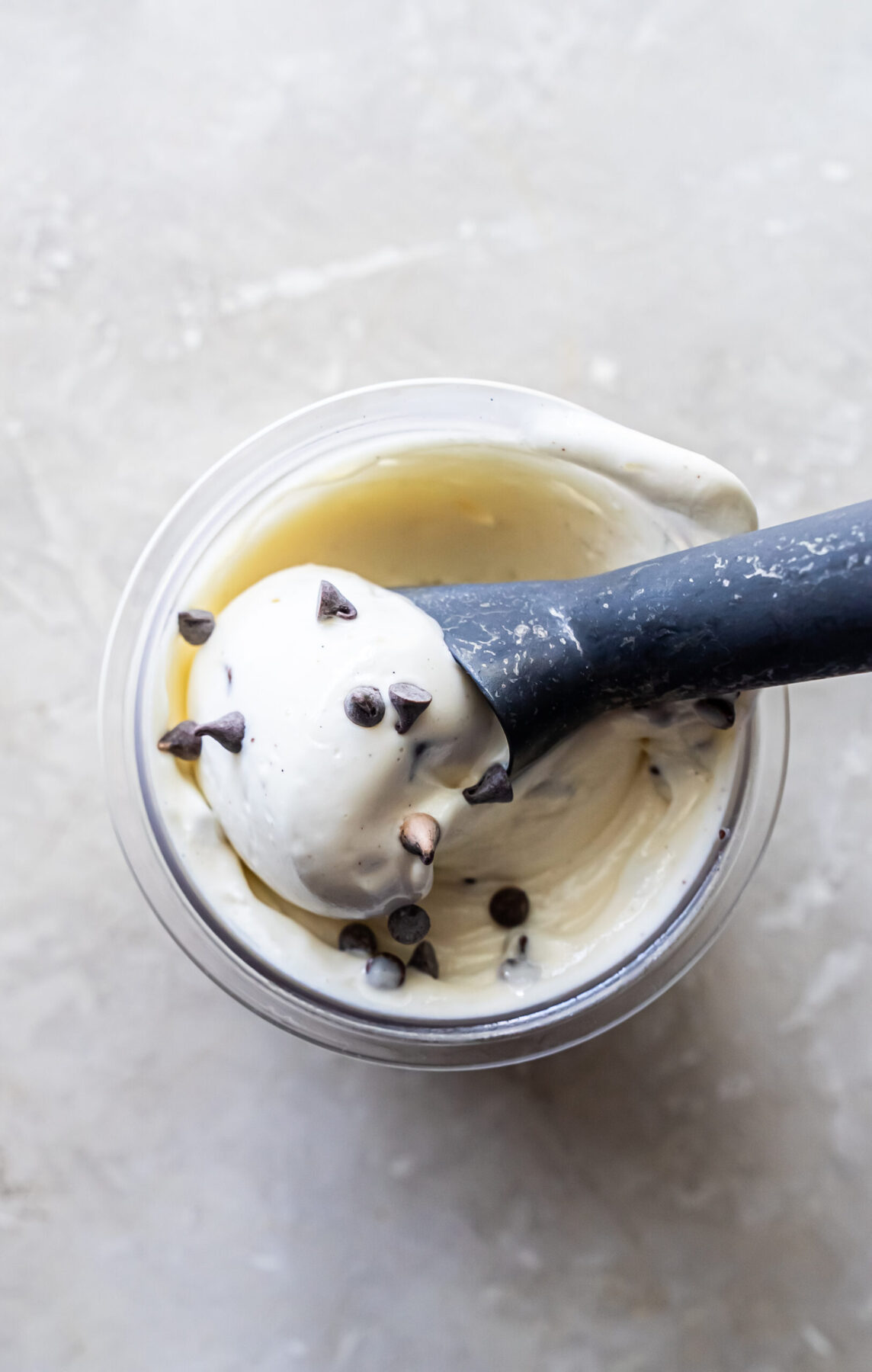 Top-down view of a black scoop holding a round portion of Mint Chocolate Protein Ice Cream over a clear pint. Mini chocolate chips are scattered on the white surface and mixed into the frozen dessert below.
