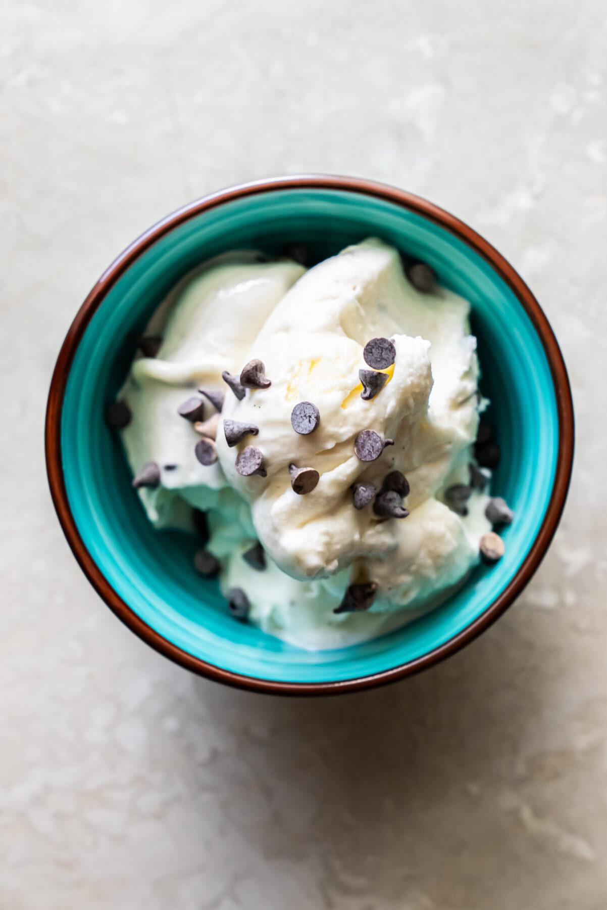 Overhead shot of scoops of Mint Chocolate Protein Ice Cream served in a teal ceramic bowl with a brown rim. The white ice cream is topped with numerous mini chocolate chips, resting on a textured grey surface.