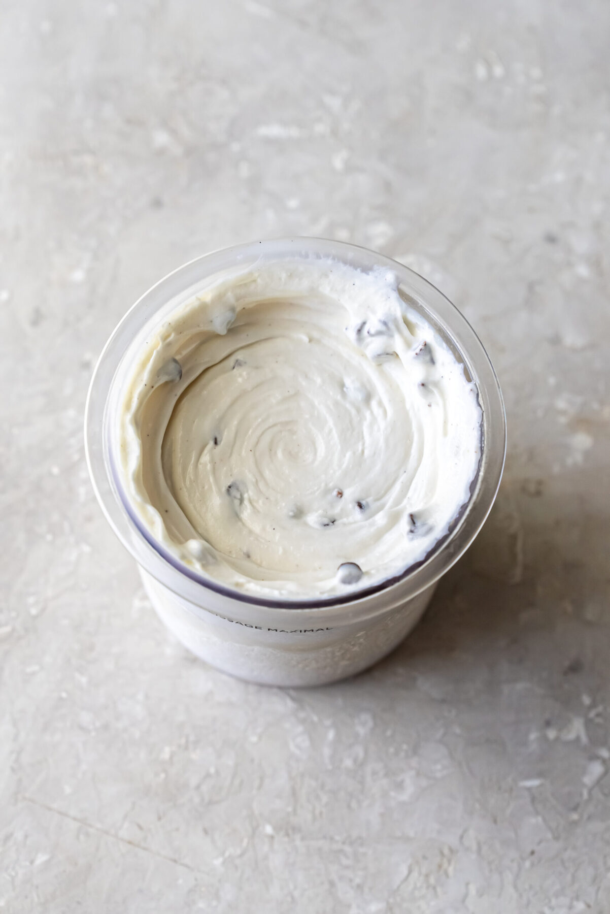 Direct top-down view of swirled Mint Chocolate Protein Ice Cream inside a clear Ninja Creami pint. Dark chocolate chips are distributed throughout the smooth white mixture, showing the texture after processing.