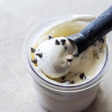 Close-up of a black ice cream scoop lifting a ball of white Mint Chocolate Protein Ice Cream from a clear Ninja Creami pint. Dark chocolate chips are scattered on top and mixed into the creamy base.
