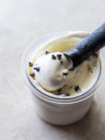 Close-up of a black ice cream scoop lifting a ball of white Mint Chocolate Protein Ice Cream from a clear Ninja Creami pint. Dark chocolate chips are scattered on top and mixed into the creamy base.