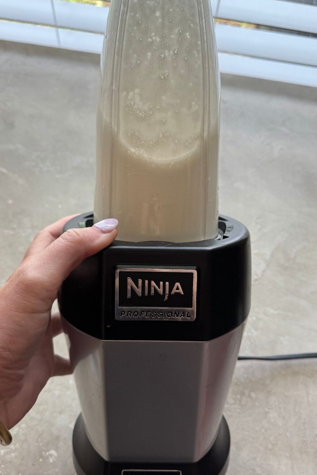 A hand holds a Ninja Professional blender cup filled with frothy white liquid base, blending ingredients for Mint Chocolate Protein Ice Cream before freezing.