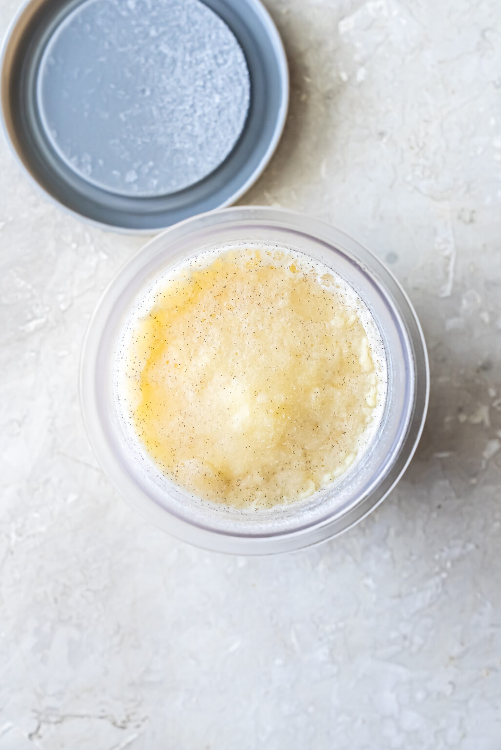 Top-down view of a frozen, pale yellow ice cream base speckled with vanilla bean seeds inside a clear Ninja Creami pint. A grey lid sits beside it on a textured surface.