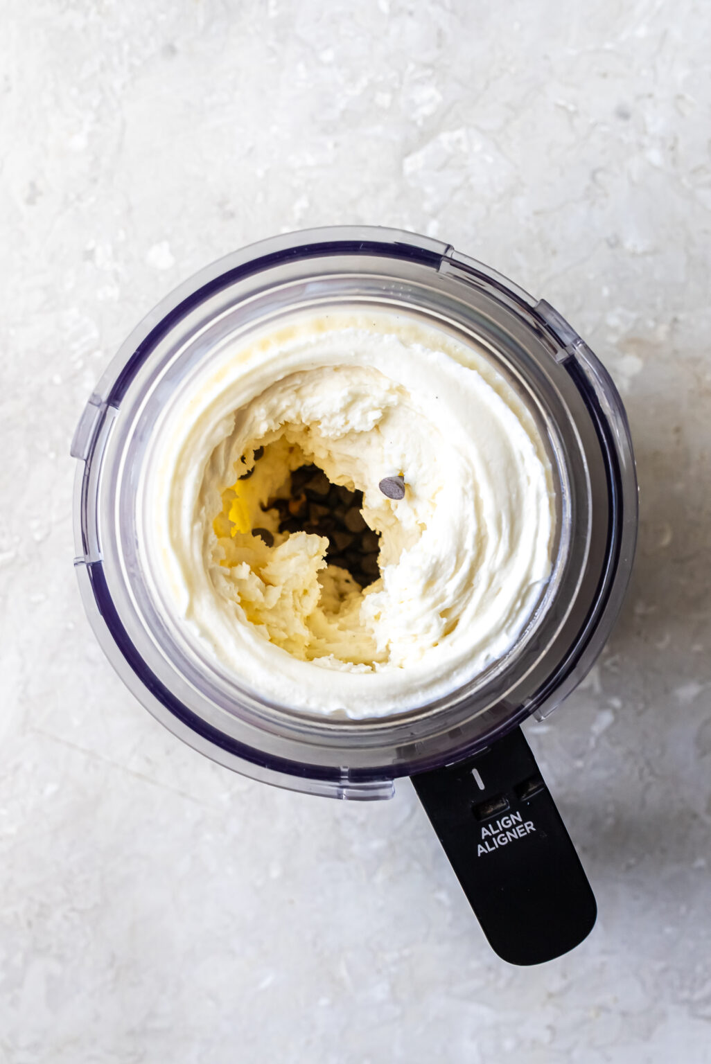 Overhead shot of mini chocolate chips poured into the center well of spun white ice cream in a Ninja Creami pint, preparing for the mix-in cycle to create Mint Chocolate Protein Ice Cream.