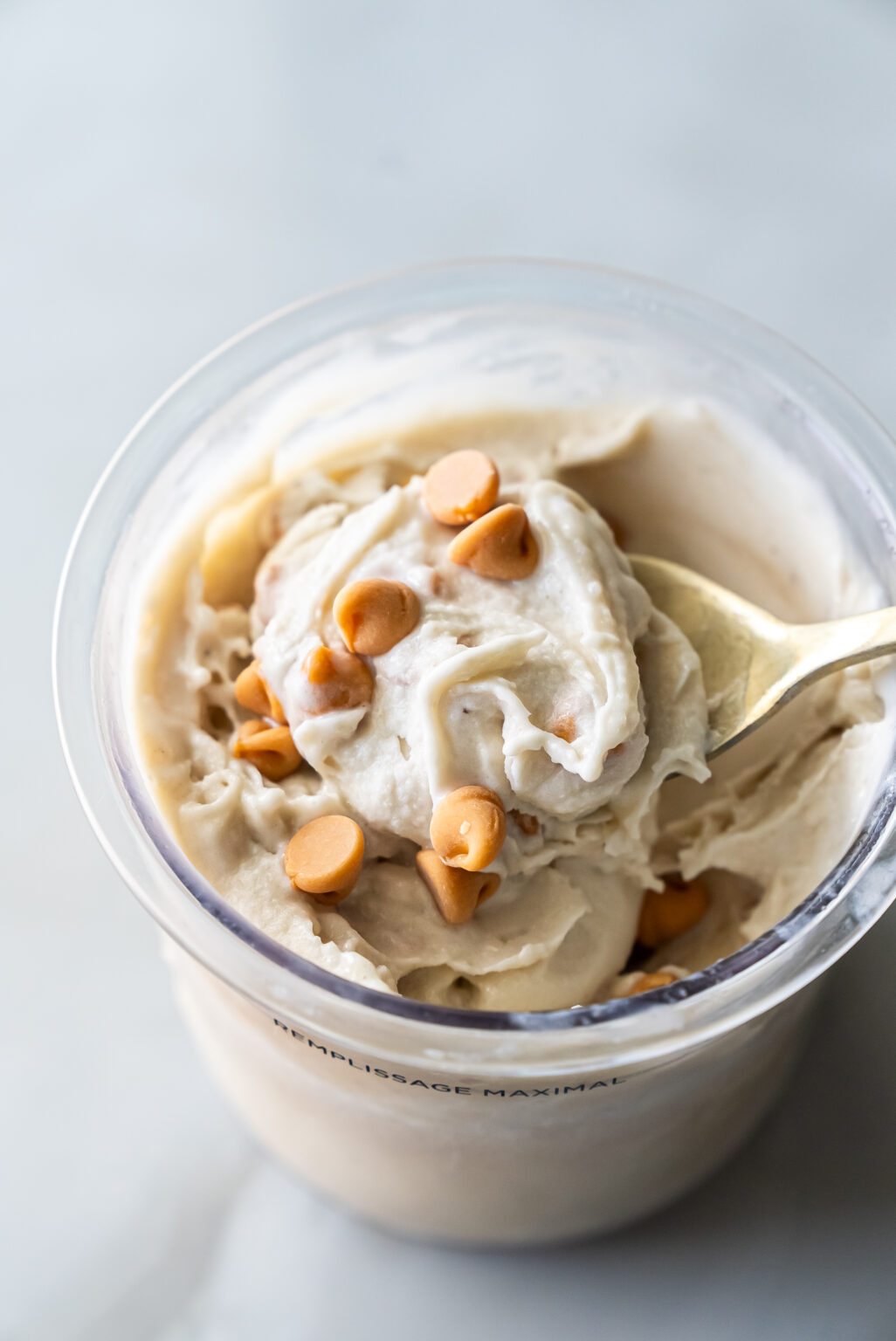 Top view of creamy Ninja Creami Banana Ice Cream with protein powder in a clear pint, topped with butterscotch chips and a gold spoon lifting a swirl, highlighting its thick texture.