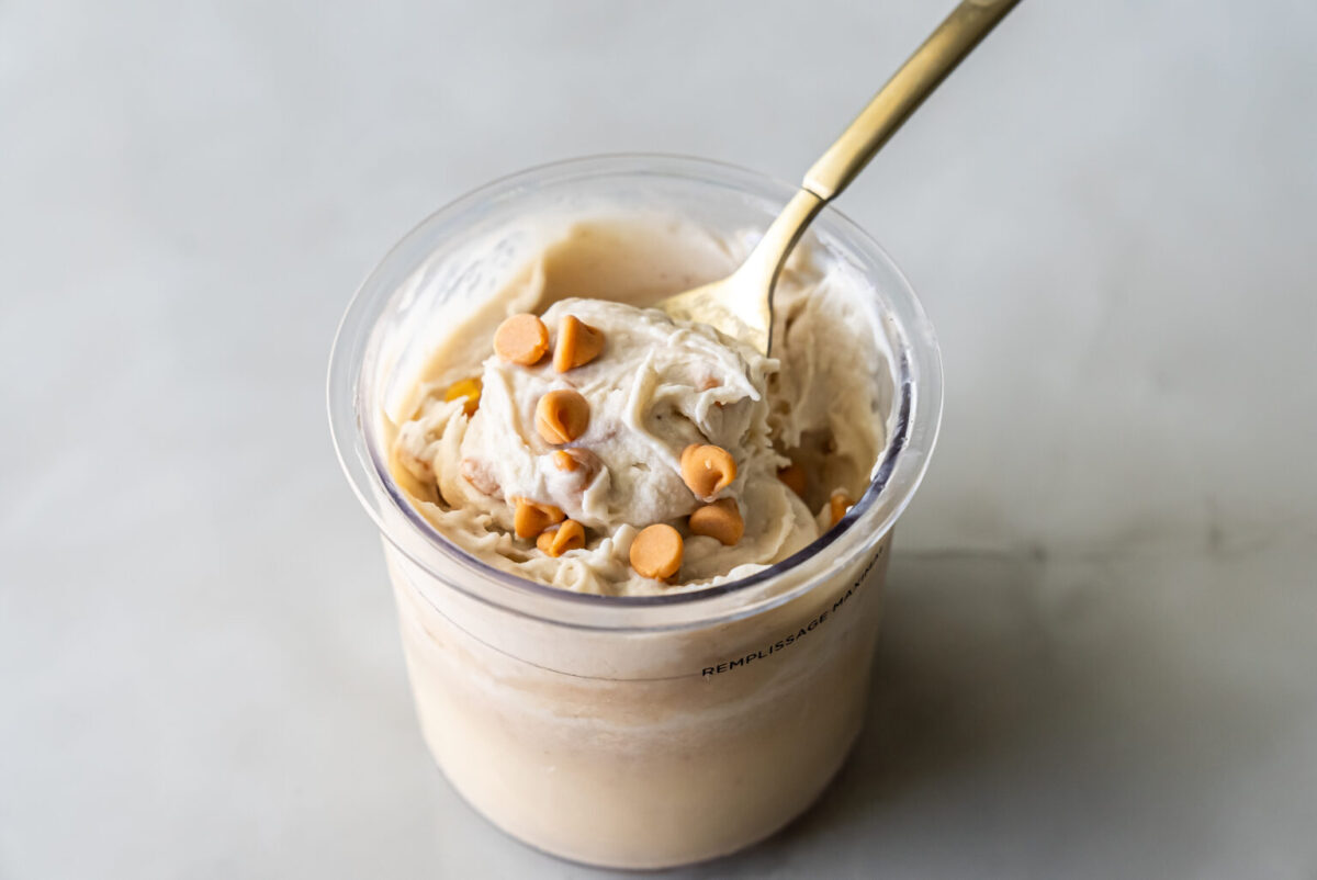 Creamy Ninja Creami Banana Ice Cream with protein powder in a clear pint, topped with butterscotch chips and a gold spoon lifting a swirl, highlighting its thick texture.