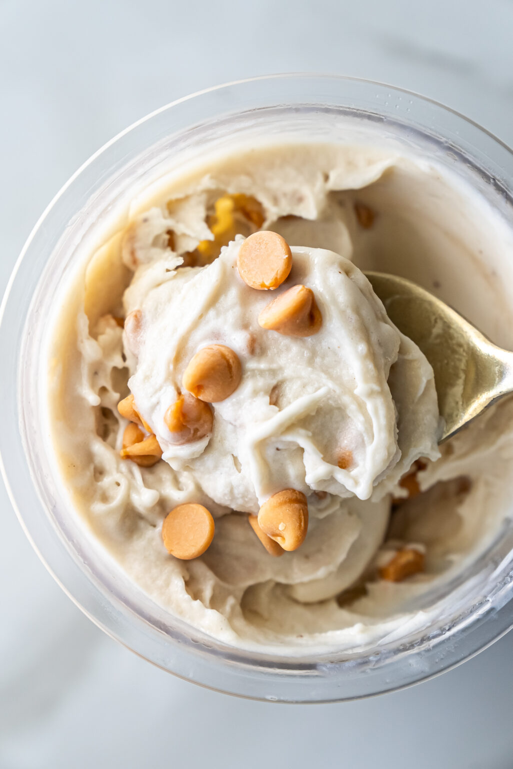 Top view of creamy Ninja Creami Banana Ice Cream with protein powder in a clear pint, topped with butterscotch chips and a gold spoon lifting a swirl, highlighting its thick texture.