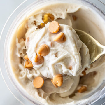Top view of creamy Ninja Creami Banana Ice Cream with protein powder in a clear pint, topped with butterscotch chips and a gold spoon lifting a swirl, highlighting its thick texture.