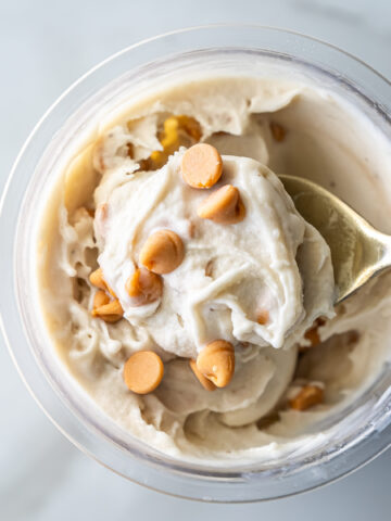 Top view of creamy Ninja Creami Banana Ice Cream with protein powder in a clear pint, topped with butterscotch chips and a gold spoon lifting a swirl, highlighting its thick texture.