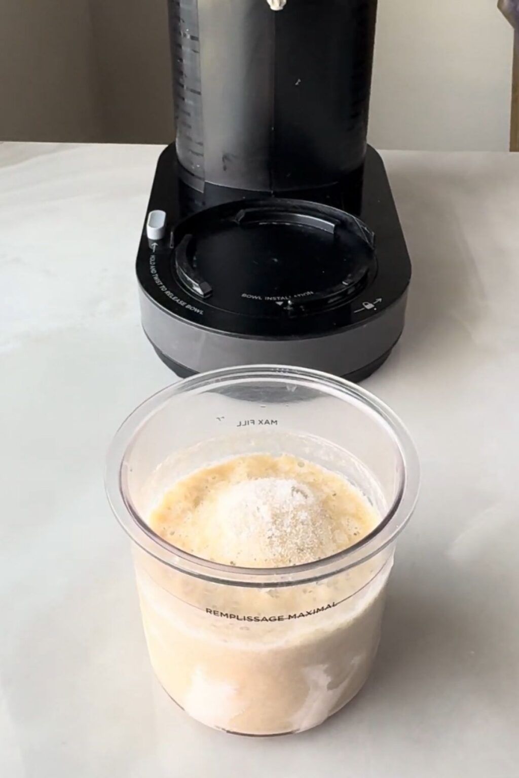 A clear Ninja Creami pint sitting on a counter next to the machine base, filled with unblended banana and protein powder mixture before processing for Ninja Creami Banana Ice Cream.