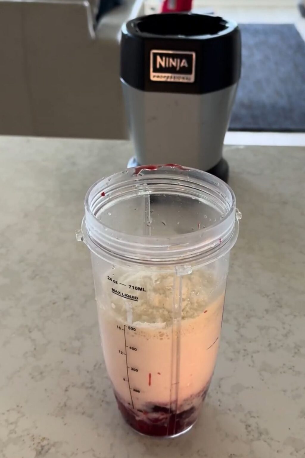 A clear Ninja blender cup on a marble countertop contains layered ingredients: dark red cherry puree at the bottom, creamy white liquid above, and powder on top—ready to blend into Ninja Creami Cherry Protein Ice Cream.