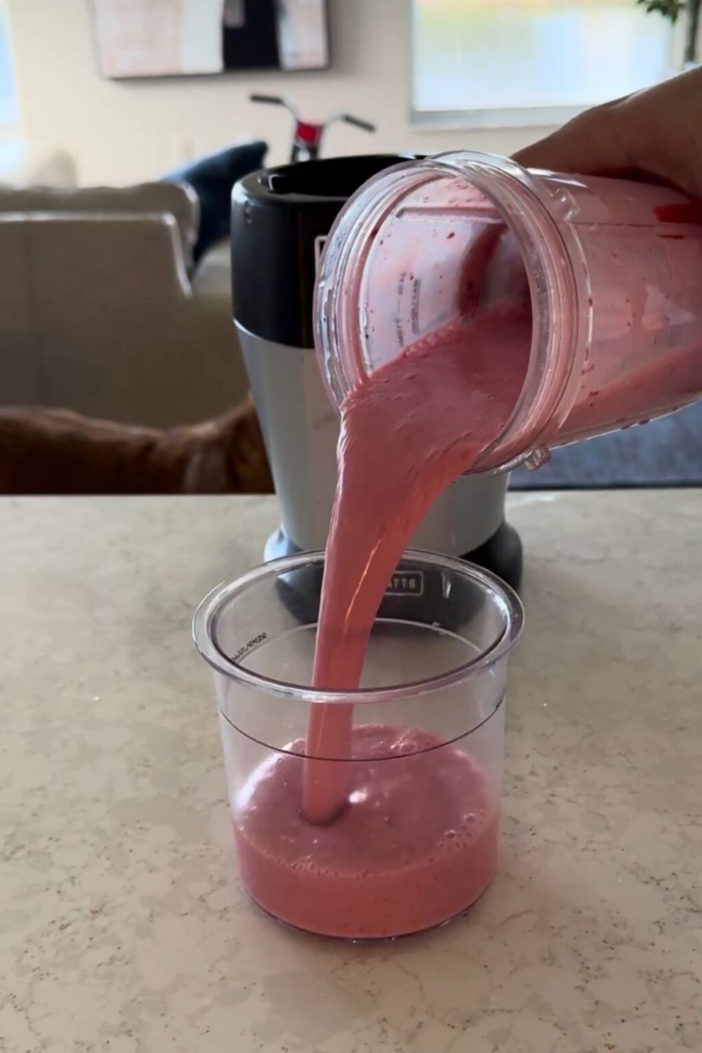 Hand pouring thick, frothy pink cherry-protein blend from blender jar into empty Ninja Creami pint, filling it precisely for freezing and later churning into Ninja Creami Cherry Protein Ice Cream.
