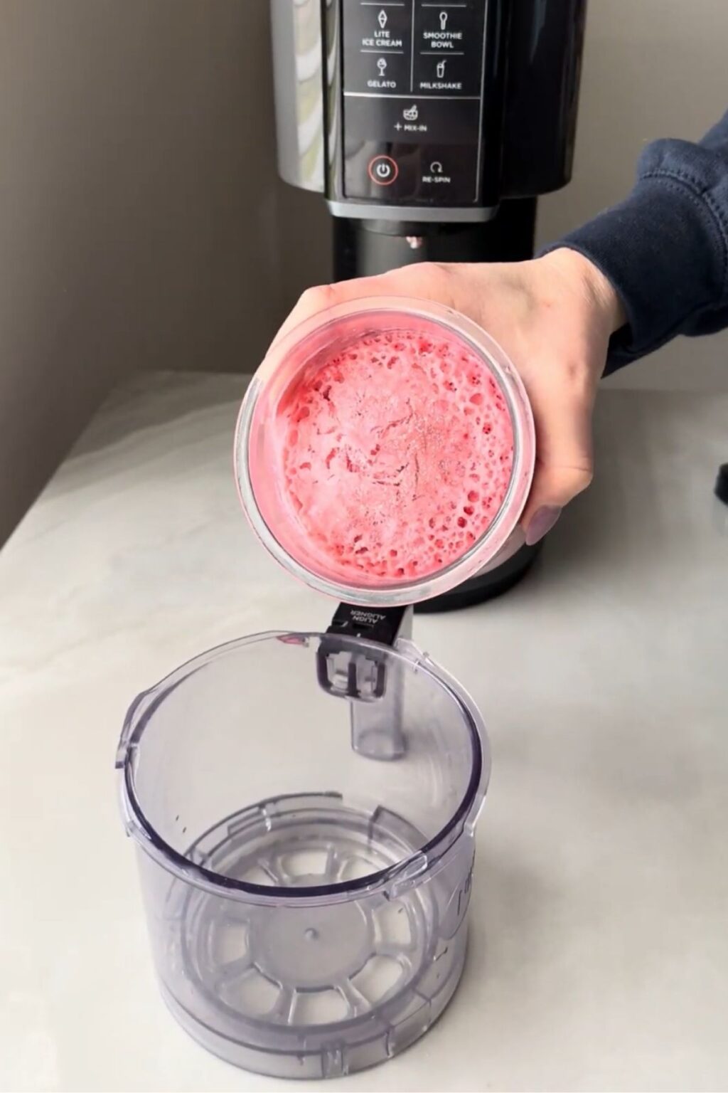 Hand holding a Ninja Creami pint containing frozen, bubbly pink cherry-protein base above an empty pint holder, preparing to insert into machine for churning into Ninja Creami Cherry Protein Ice Cream.