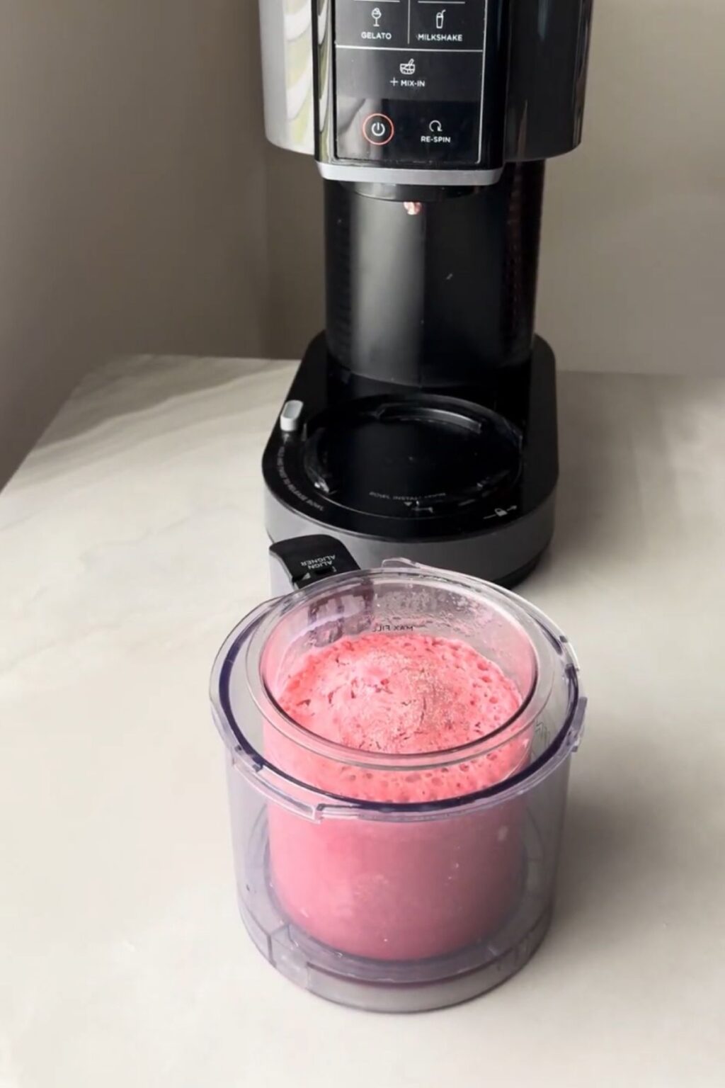 Clear Ninja Creami pint filled with frozen, aerated pink cherry-protein base, placed on counter next to machine — prepped for spinning into creamy Ninja Creami Cherry Protein Ice Cream.