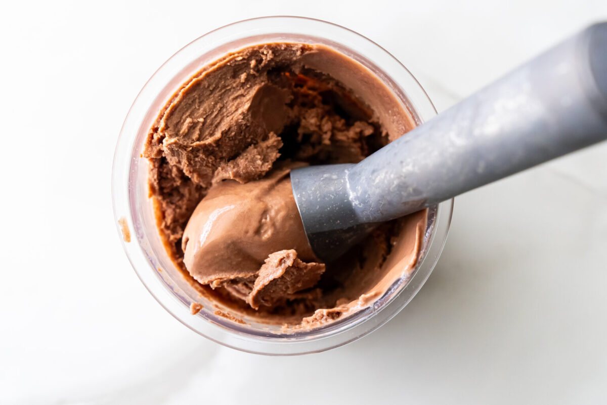 Top-down view of rich, creamy Ninja Creami Chocolate Malt Ice Cream in a clear pint, with a gray ice cream scoop lifting a smooth swirl, showcasing its dense, fudgy texture after churning.