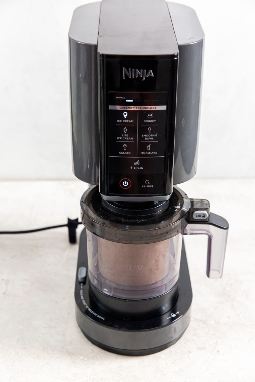 Front view of a black Ninja Creami machine with display showing settings like Ice Cream and Gelato, actively processing a pint of chocolate malt base into creamy Ninja Creami Chocolate Malt Ice Cream.