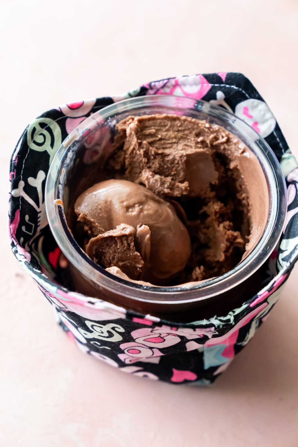 A clear pint of Ninja Creami Chocolate Malt Ice Cream nestled in a colorful patterned fabric cozy, featuring a perfect scoop on top, highlighting its indulgent, homemade appeal against a soft pink background.