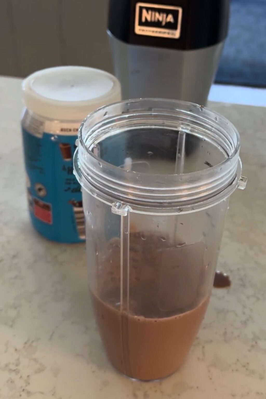 A clear Ninja blender cup filled with a smooth, light brown chocolate malt liquid base sits on a marble countertop, next to a tub of malt powder and a Ninja Professional blender.