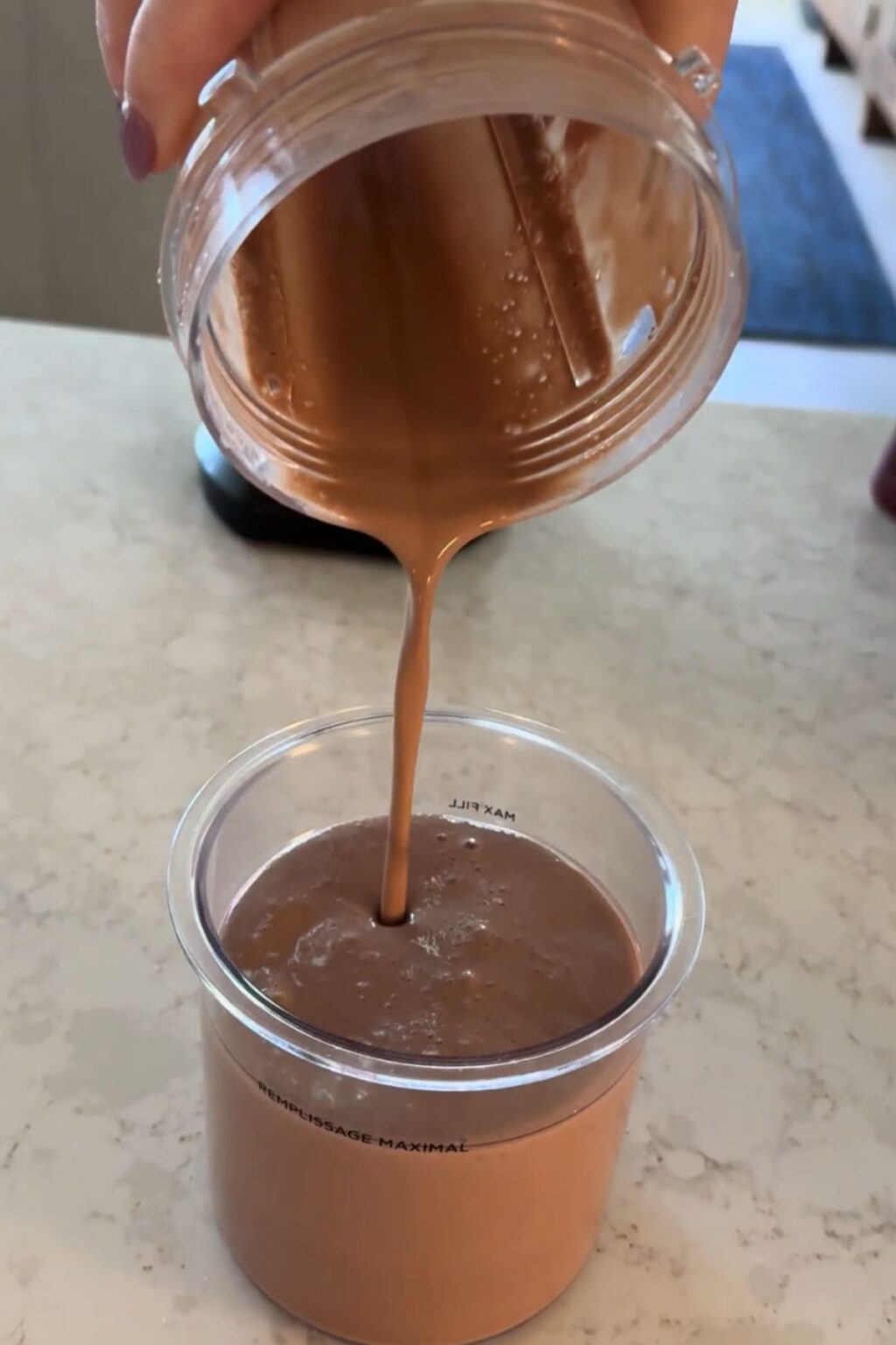 A hand pours smooth, thick chocolate malt liquid from a blender jar into a clear Ninja Creami pint marked “MAX FILL,” filling it precisely for freezing and later churning into Ninja Creami Chocolate Malt Ice Cream.