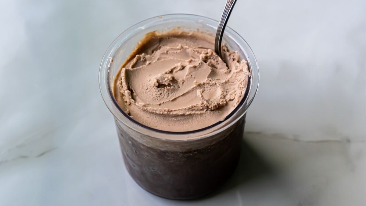 A clear plastic container filled with dairy-free chocolate ice cream and a spoon placed inside, set on a light-colored marble surface.