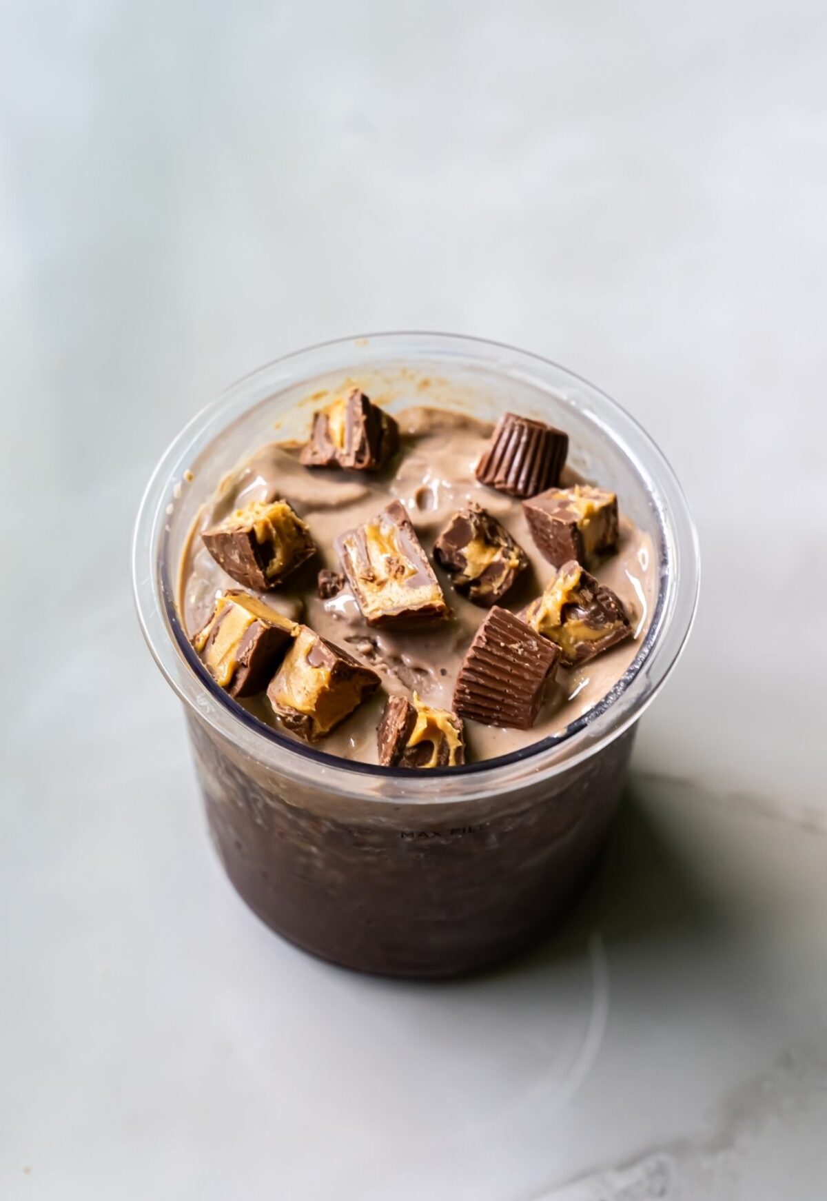 A cup filled with dairy-free chocolate and peanut butter dessert, topped with chunks of peanut butter cups, placed on a light gray surface. Perfectly crafted using the Ninja Creami, this treat is also a protein ice cream delight.