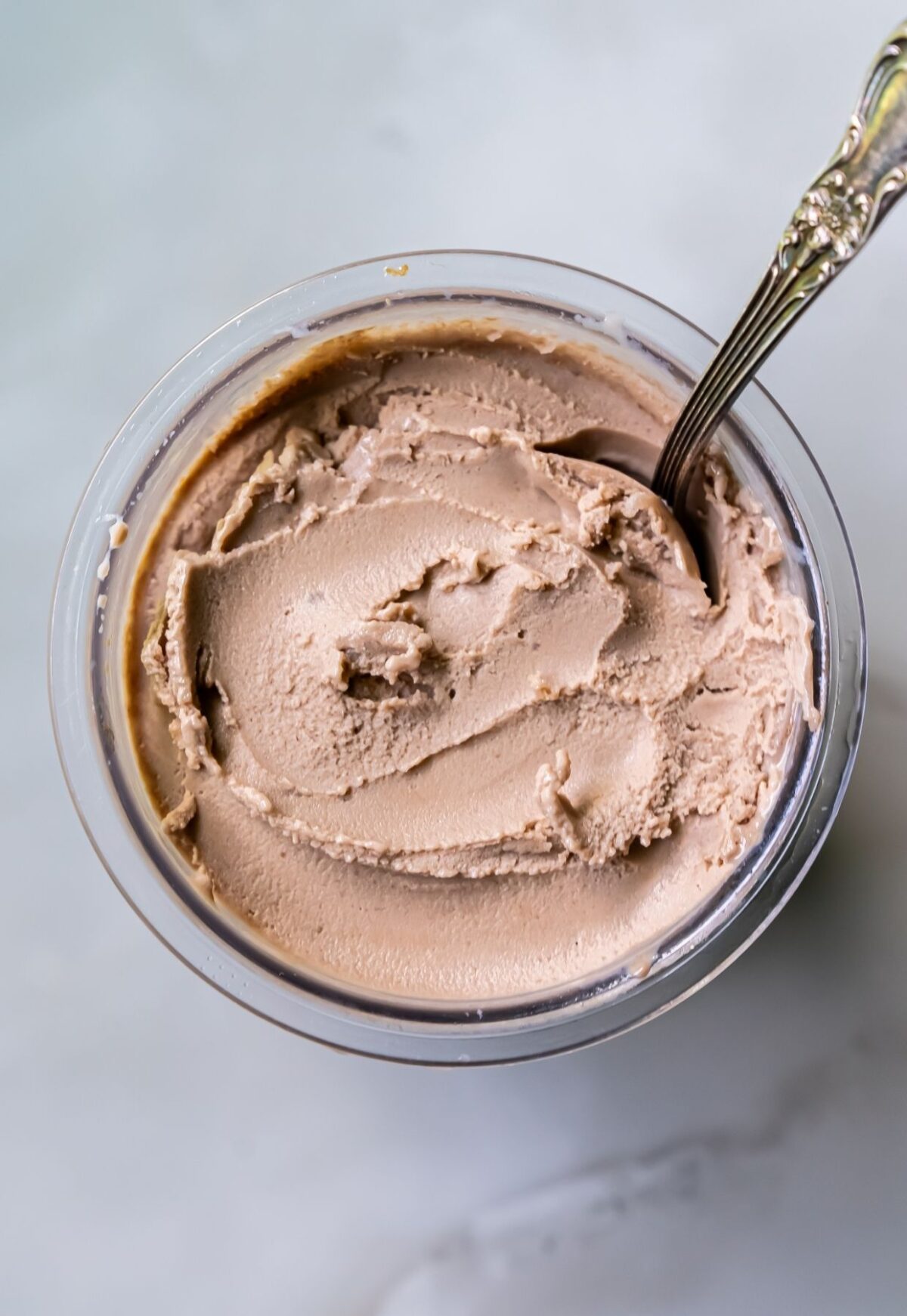 A close-up image of a container filled with brown, creamy, and slightly textured protein ice cream, with a metal spoon inserted into it.