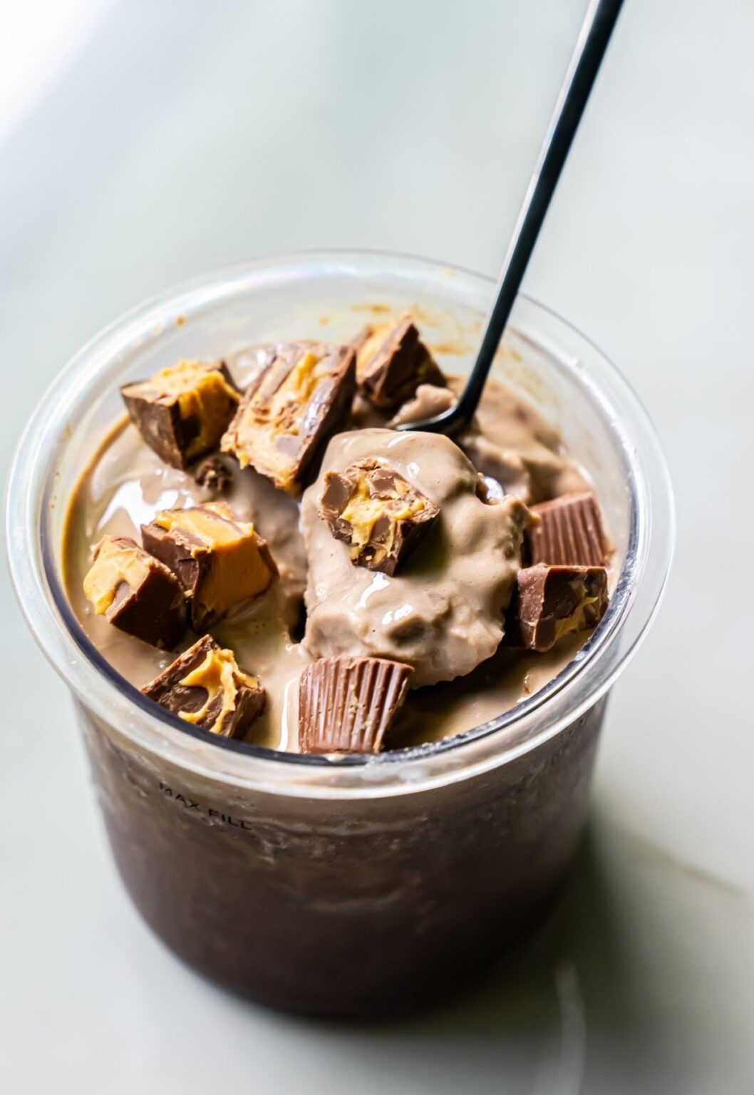 Top view of high protein chocolate ice cream in a clear pint, topped with chopped peanut butter cups and a black spoon.