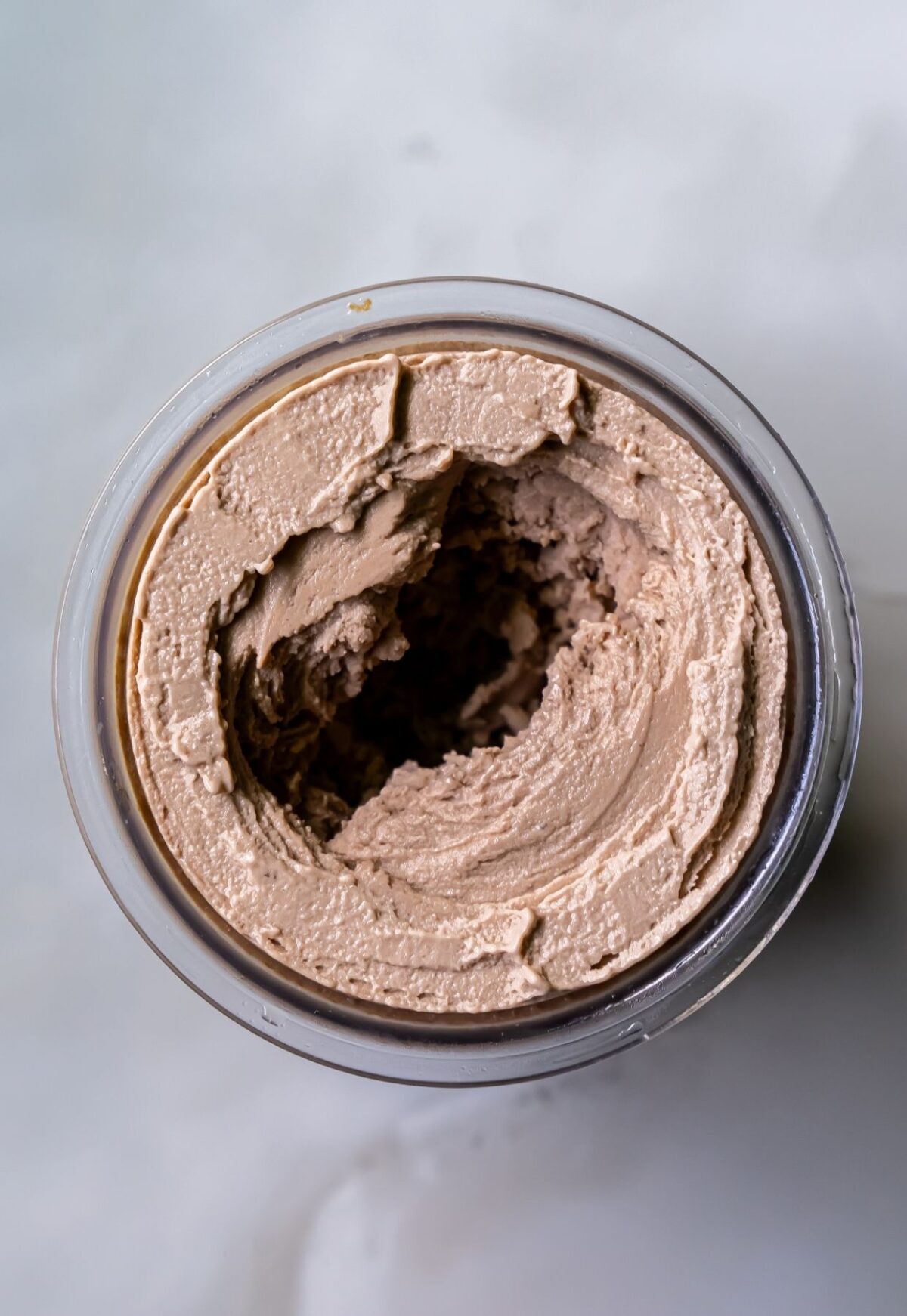 Top view of an opened container of light brown, creamy spread, which appears to be partially used and has a scoop taken from the center. Resembling chocolate peanut butter protein ice cream, the container is placed on a light-colored surface.
