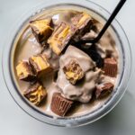 A glass bowl filled with dairy-free, protein ice cream is topped with chunks of chocolate and peanut butter candy, while a black spoon rests inside.