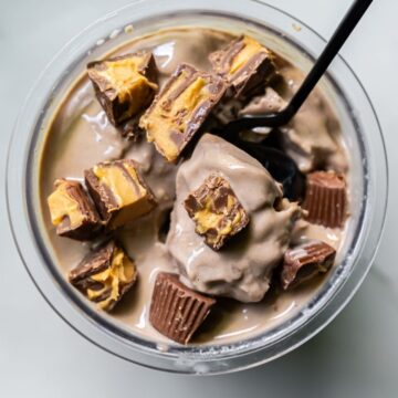 A glass bowl filled with dairy-free, protein ice cream is topped with chunks of chocolate and peanut butter candy, while a black spoon rests inside.
