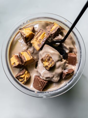 A glass bowl filled with dairy-free, protein ice cream is topped with chunks of chocolate and peanut butter candy, while a black spoon rests inside.