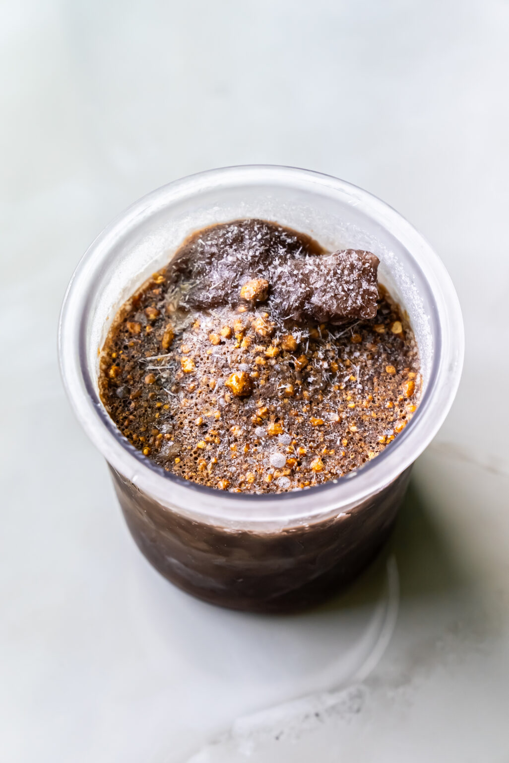 A close-up of a small glass container filled with a rich, dairy-free dark chocolate dessert topped with crushed nuts and a dusting of powdered sugar.
