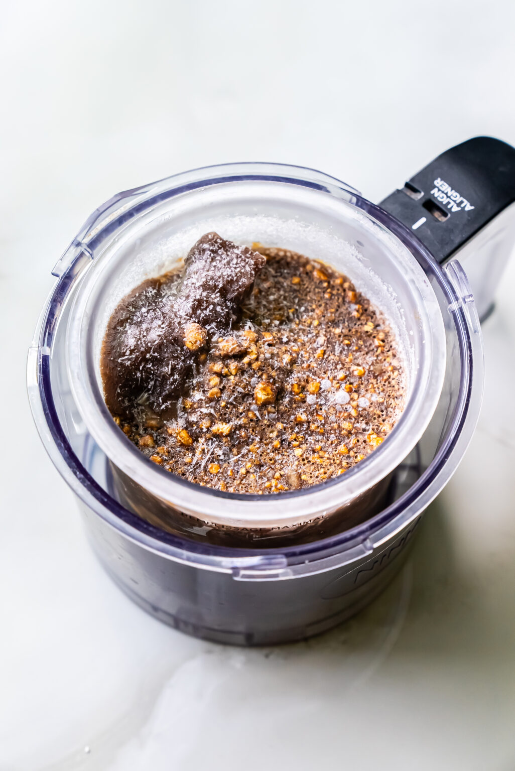 Image of a food processor filled with partially blended ingredients, including crushed ice and powdery substances, viewed from above. This dairy-free blend is perfect for creating your own homemade protein ice cream with the Ninja Creami.