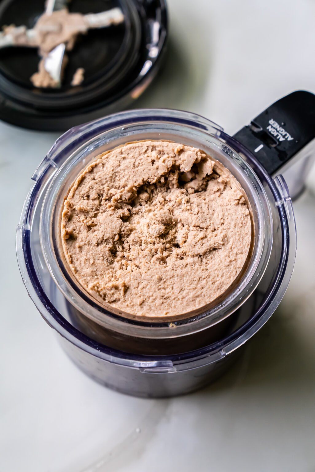 A round food processor container filled with a beige, chunky paste sits next to its lid on a white surface, resembling the beginnings of a delicious dairy-free chocolate peanut butter treat.