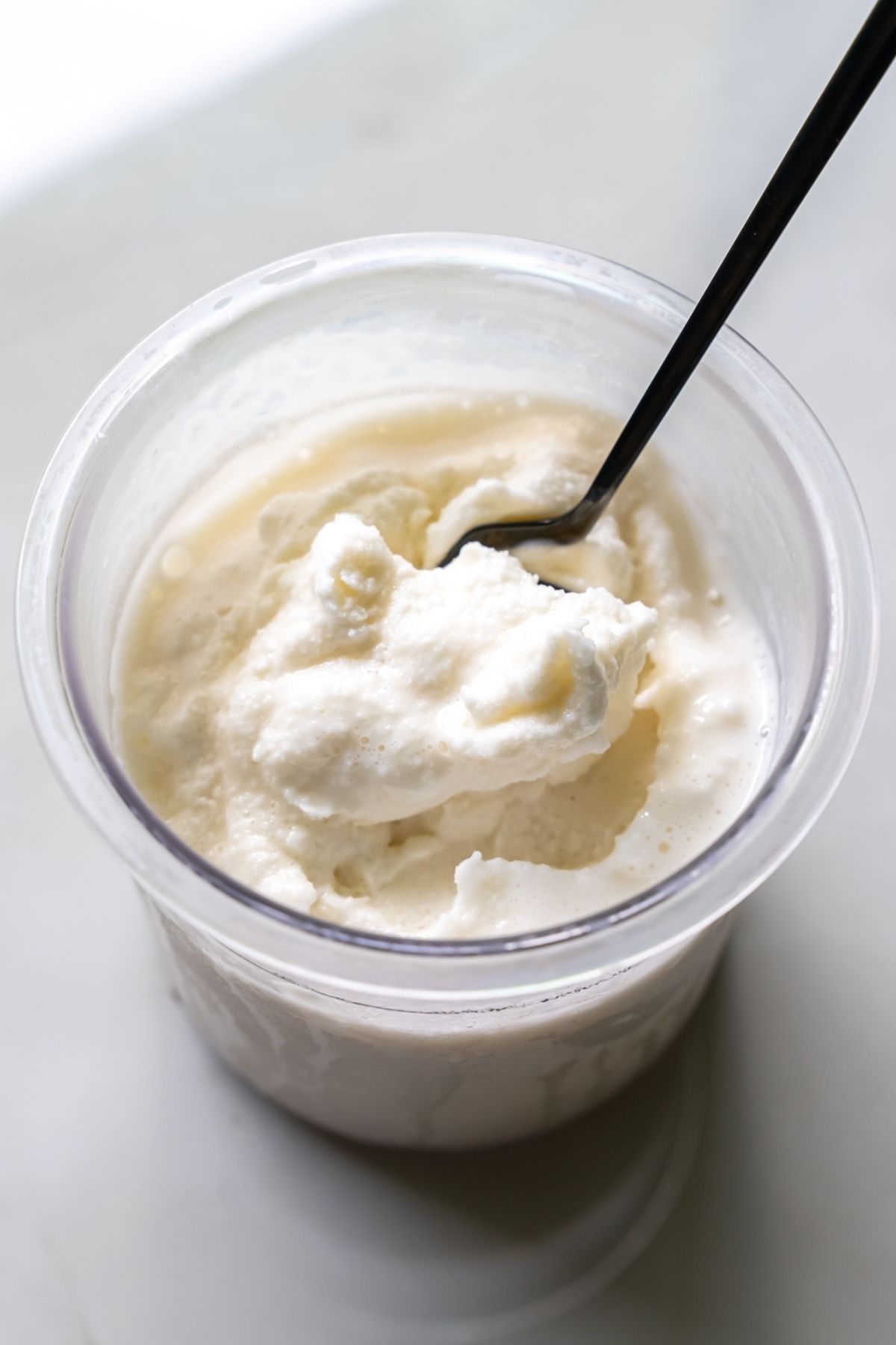 A plastic container filled with creamy white, keto-friendly ice cream, a spoon inserted into it.
