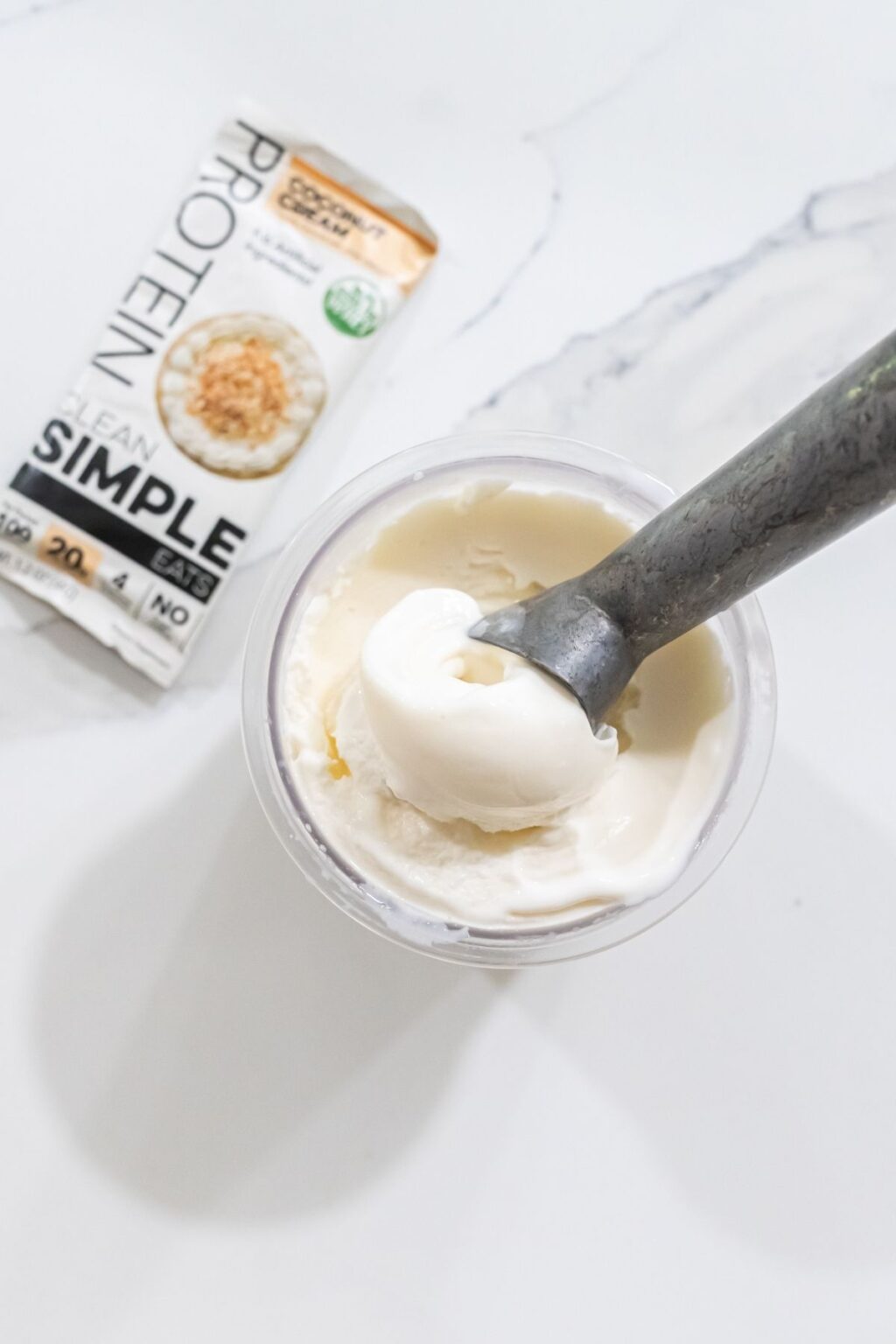 A metal scoop is inserted into a container of protein ice cream. A package of protein oats is lying beside the keto-friendly treat on a marble surface.