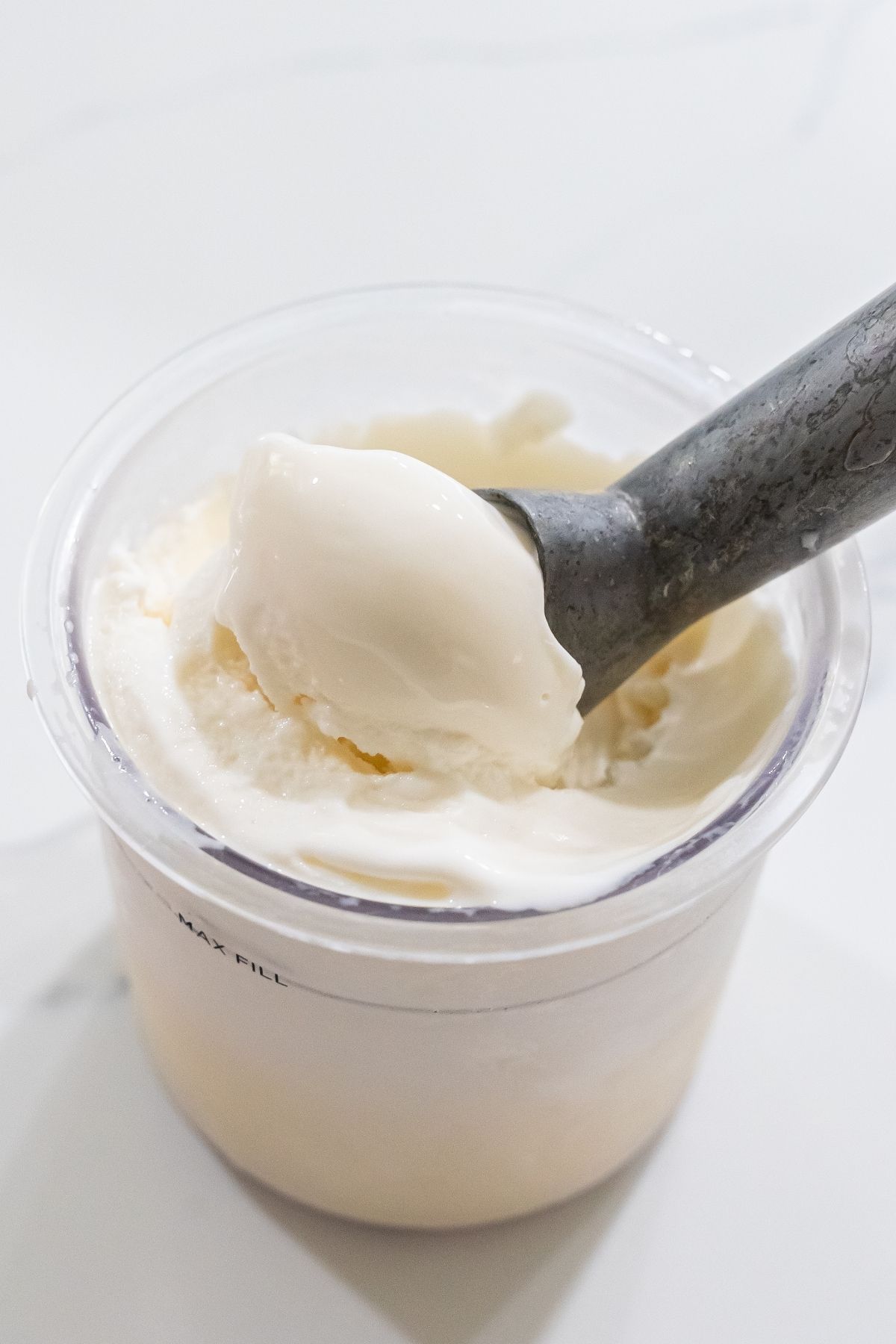 A scoop of vanilla protein ice cream is being served from a transparent container using a metal ice cream scoop.