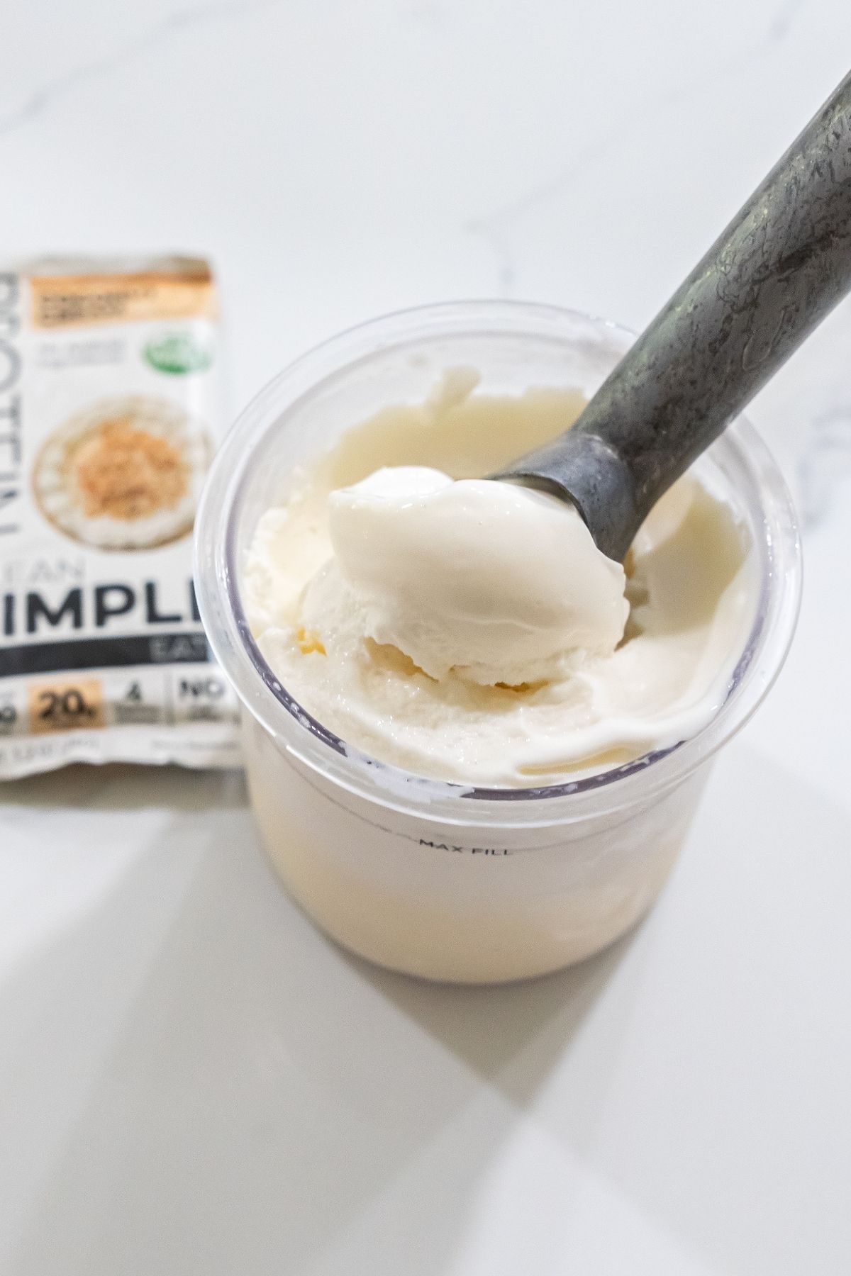 A metal spoon scoops protein ice cream from a container, resting on a white surface. A white and orange packaged protein bar is seen in the background, hinting at a delicious keto-friendly treat.
