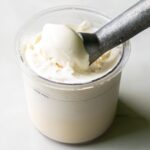 A metal scoop rests in a clear plastic container filled with Keto-friendly vanilla ice cream.