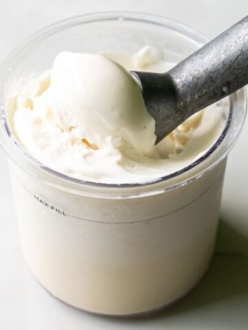 A metal scoop rests in a clear plastic container filled with Keto-friendly vanilla ice cream.