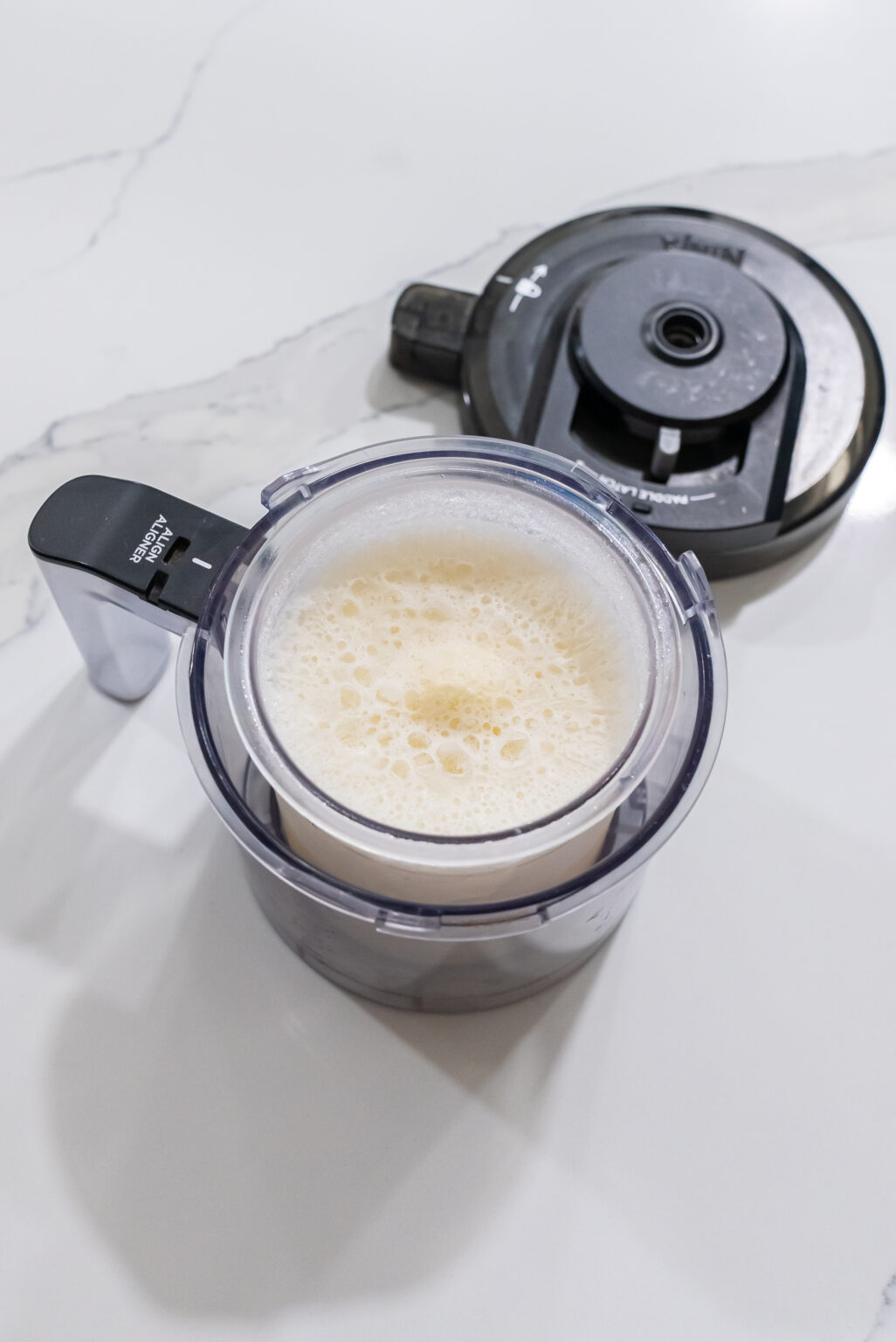 A close-up view of a Ninja Creami blender filled with frothy liquid, perfect for whipping up keto-friendly protein ice cream, with its lid placed beside it on a white surface.
