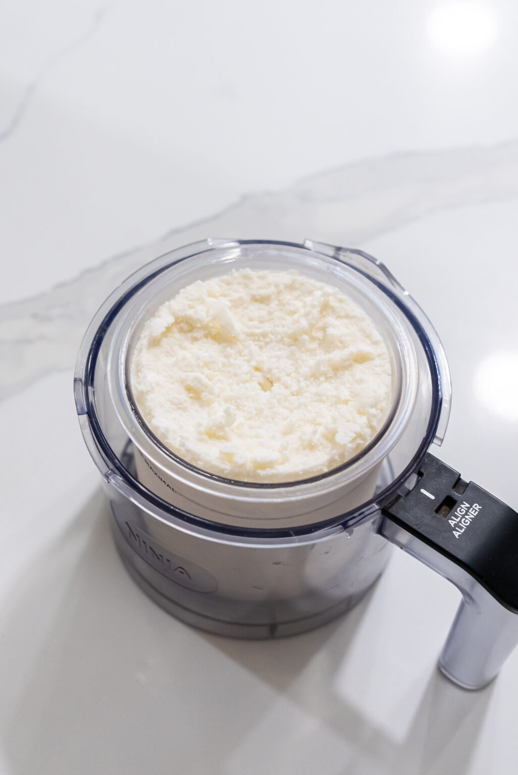 A clear plastic food processor bowl filled with shredded coconut sits on a white countertop, ready to become the base for delicious keto treats using your Ninja Creami.