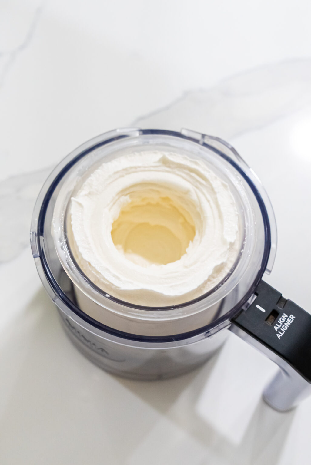 An electric Ninja Creami ice cream maker sits on a white countertop, its transparent container filled with freshly churned vanilla ice cream.