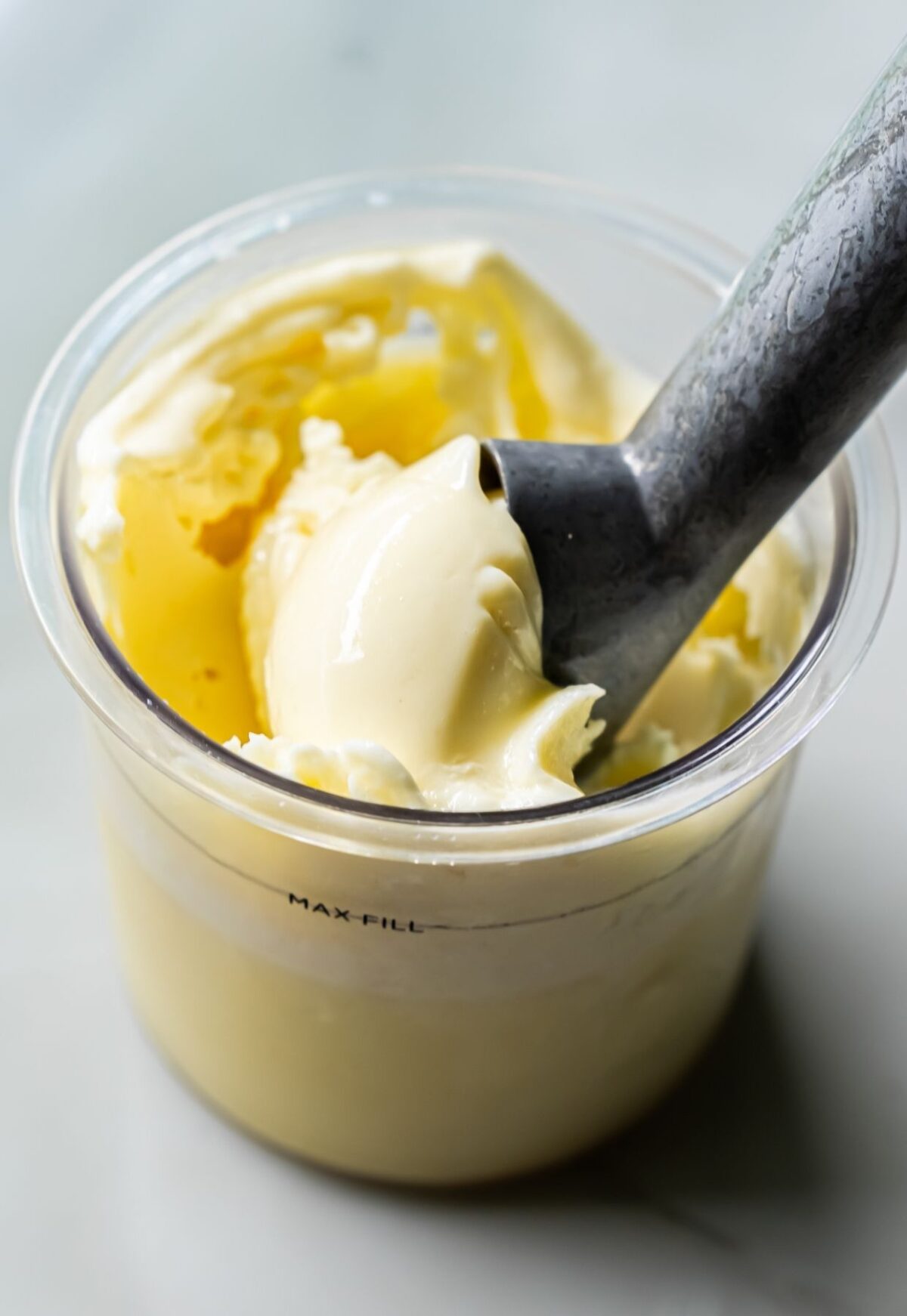 A stainless steel ice cream scoop rests in a clear container of creamy, pale yellow, low-carb protein ice cream.