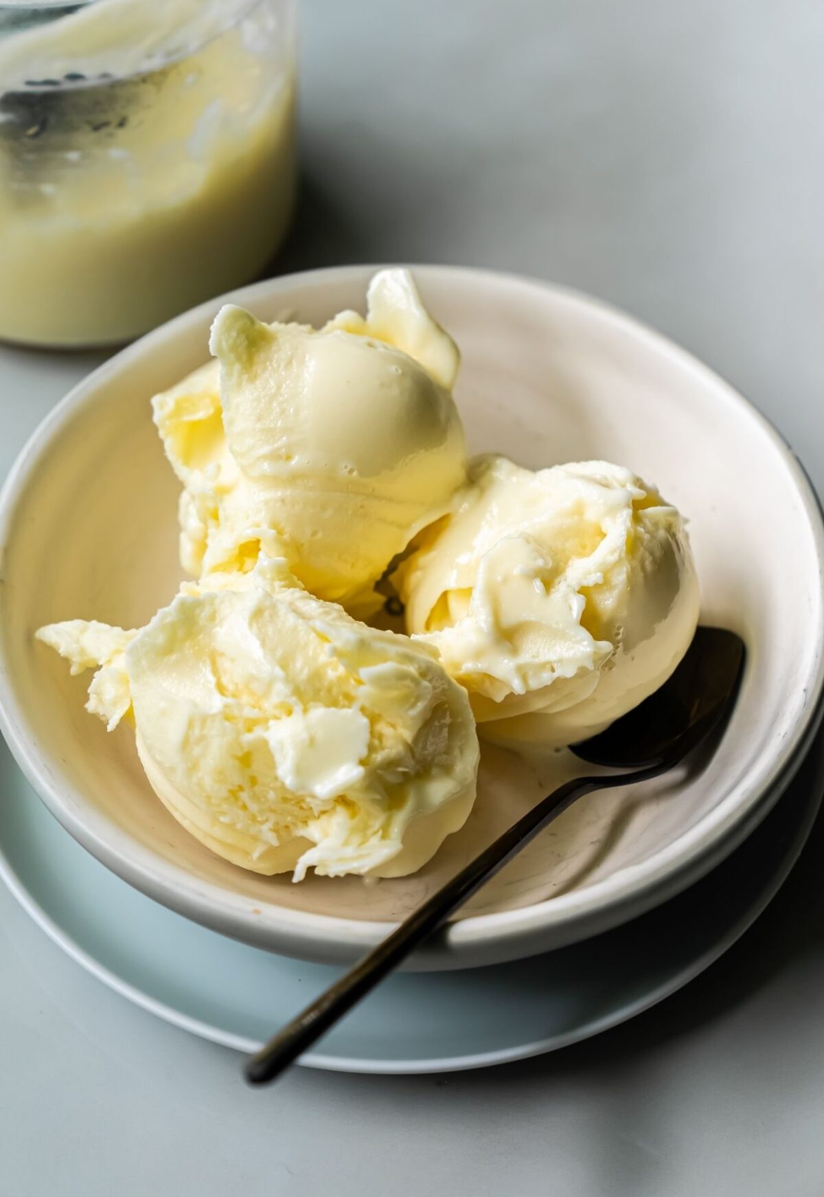 A white bowl contains three scoops of low carb, protein ice cream, accompanied by a metal spoon. A jar with a small amount of a white substance is visible in the background. Made with the Ninja Creami, this dessert is both delicious and nutritious.