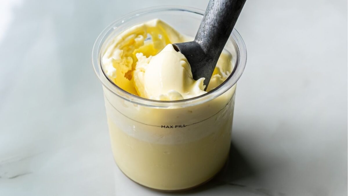 A metal ice cream scoop in a container of low carb vanilla ice cream on a light gray surface.
