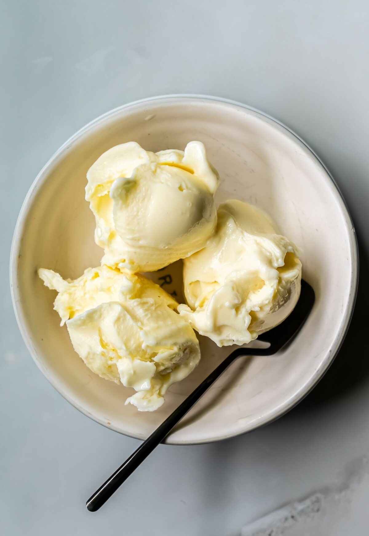 A white bowl containing three scoops of low-carb vanilla ice cream made with Ninja Creami, paired with a black spoon on a light gray surface.